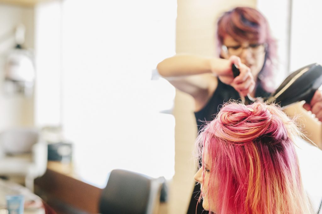 A hair stylist blowdrying a client's long pink dyed hair.